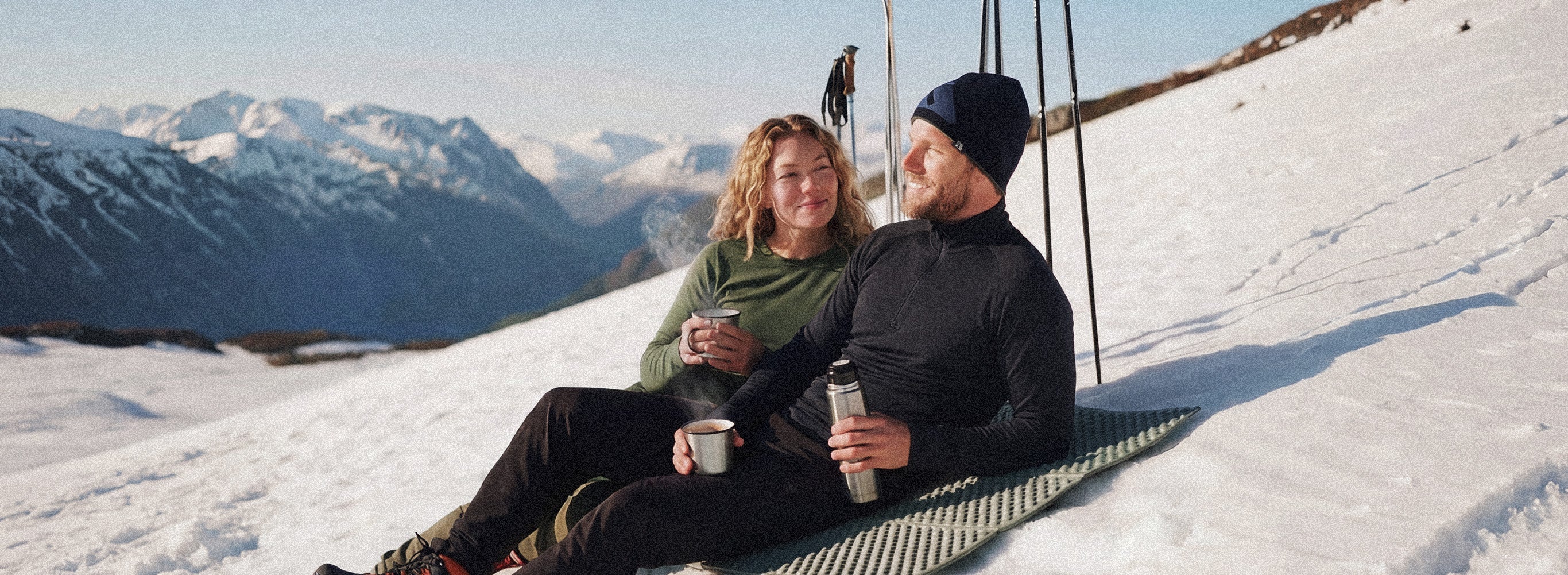 A couple sits on a mat in the snow, smiling and drinking from mugs, surrounded by snowy mountains under a clear sky. They have hiking poles and winter clothing.