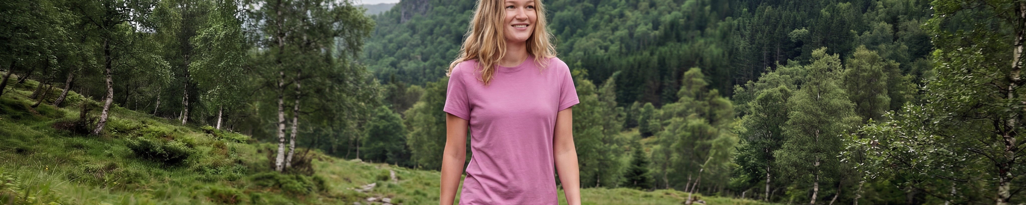 A woman with blonde hair wearing a pink T-shirt walks outdoors on a grassy, tree-lined path with green hills and forest in the background.