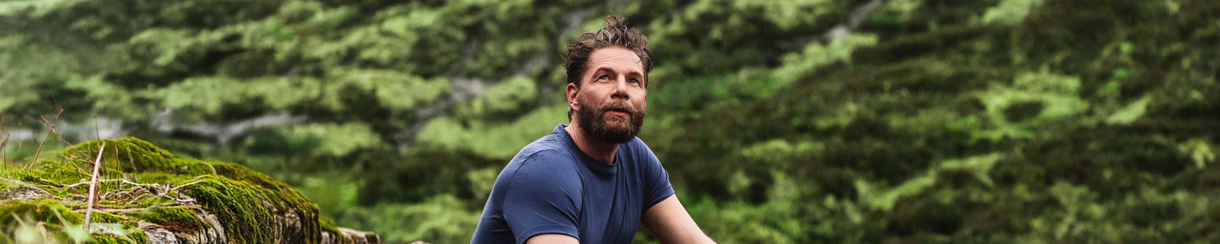 A man with tousled hair and a beard, wearing a blue t-shirt, sits outdoors on mossy rocks with a lush, green landscape in the background, looking thoughtfully into the distance.