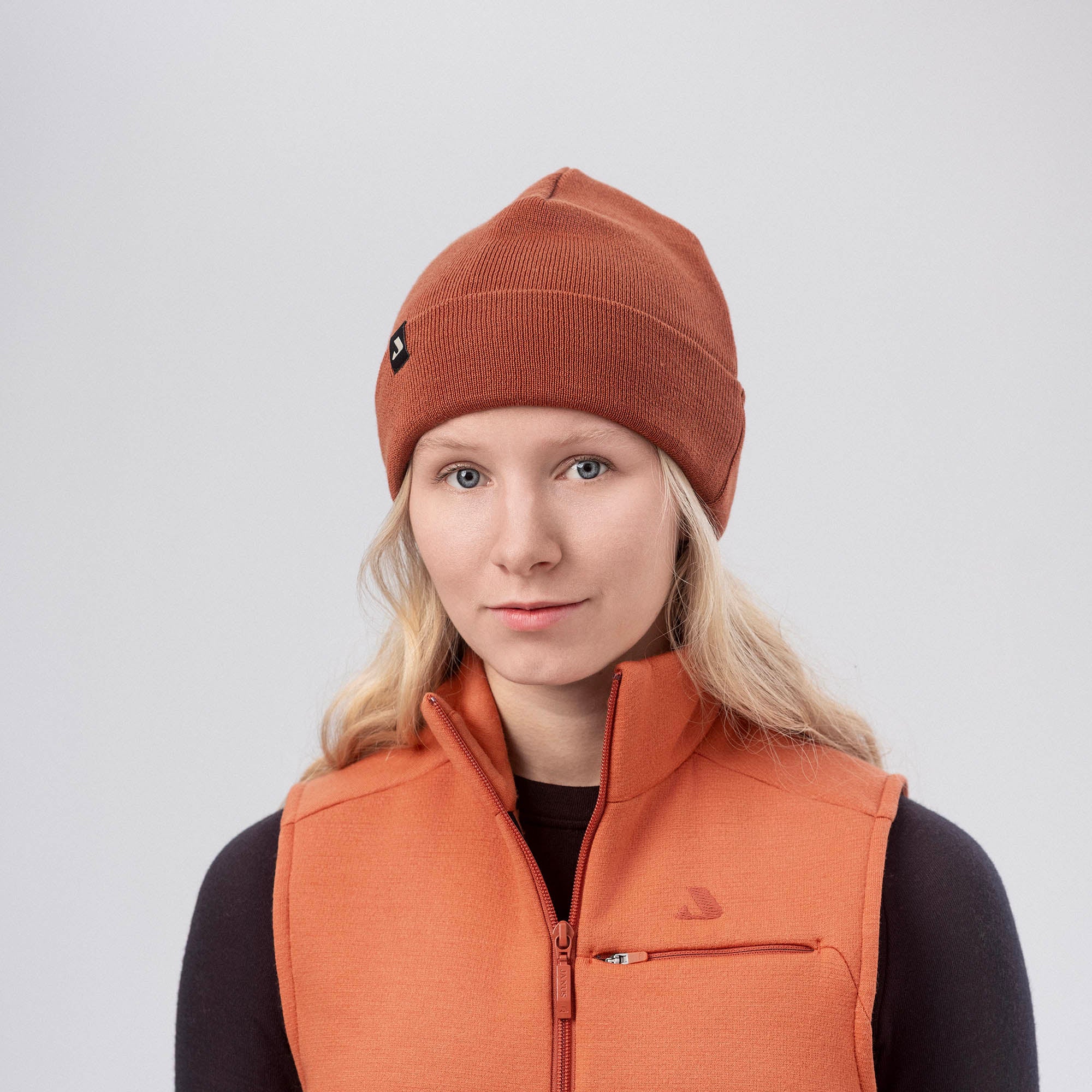 A young woman with long blonde hair wears a warm orange MeshWool beanie and an orange zip-up vest over a black top, standing against a plain light gray background.