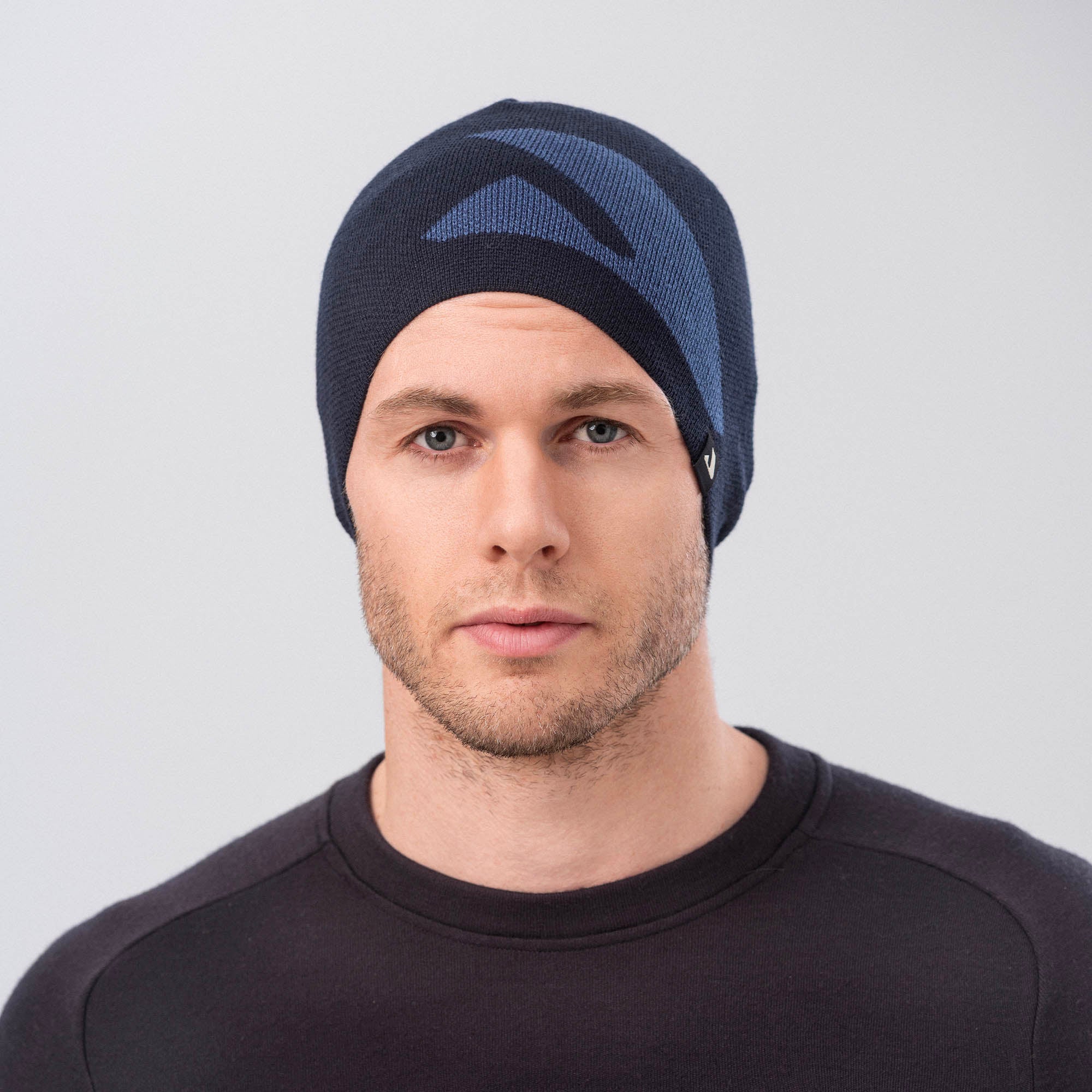 A man with light skin and short facial hair wears the dark blue Beanie – Logo for warmth and breathability, paired with a black crew-neck shirt, looking directly at the camera against a plain light background.