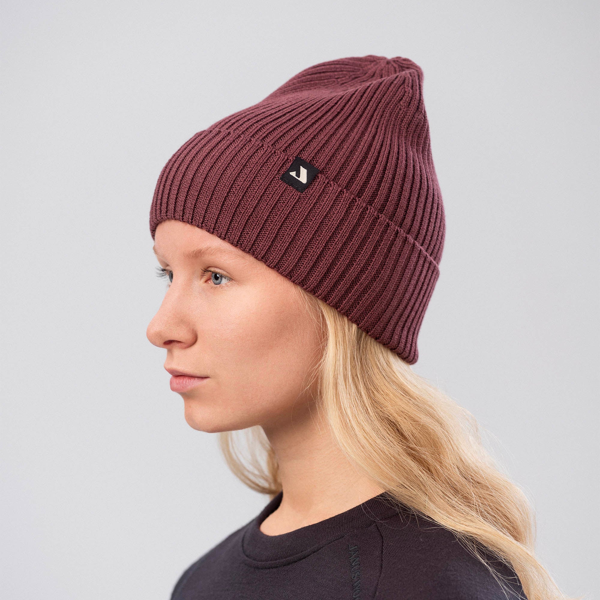 A person with long blonde hair wears the Classic Beanie in maroon, known for warmth and breathability, along with a dark top, looking to the left against a plain light gray background.