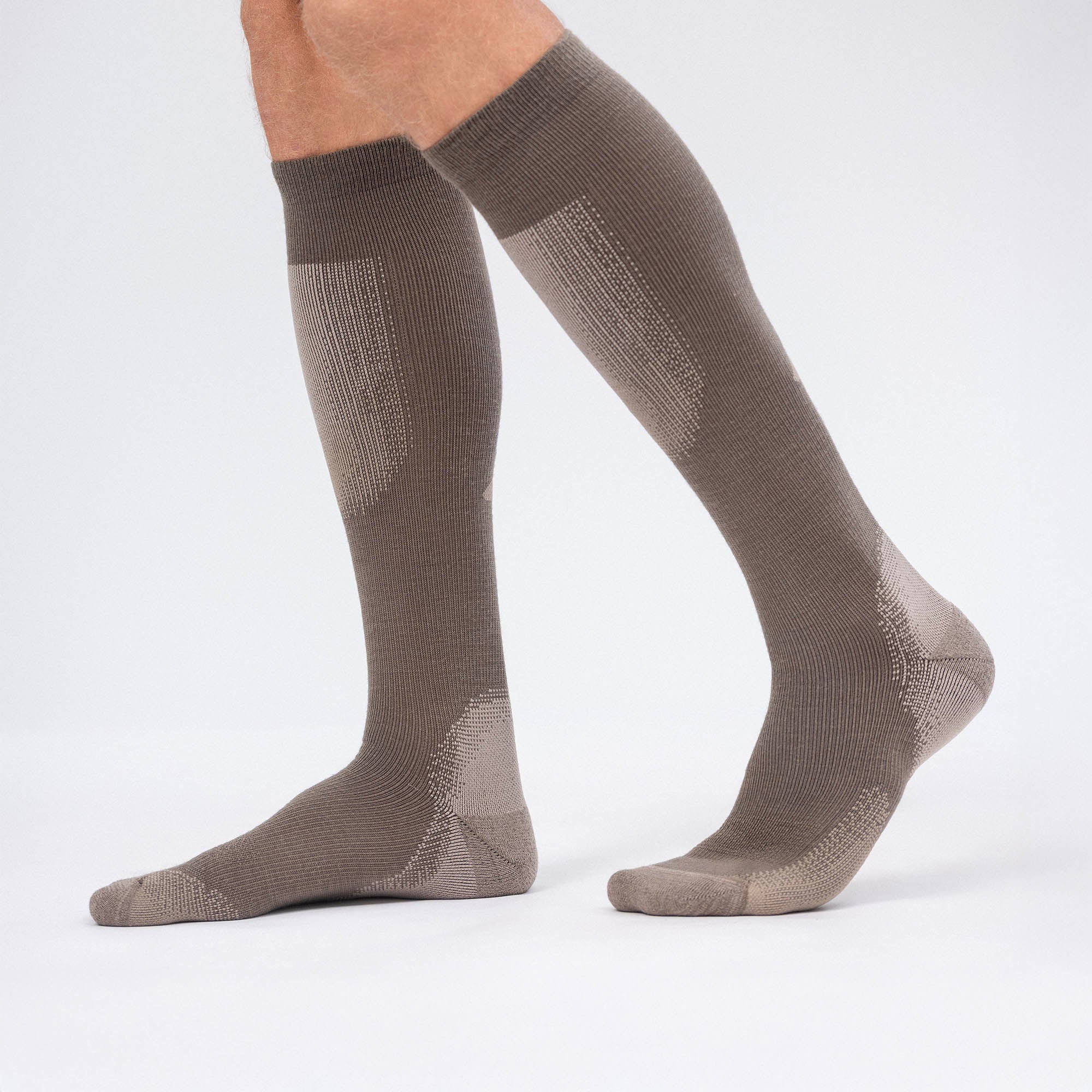A person models knee-high, light brown Socks Merino – Cross Country on a white background, with one foot flat and the other heel raised to emphasize their breathable design—perfect for cross country skiing.