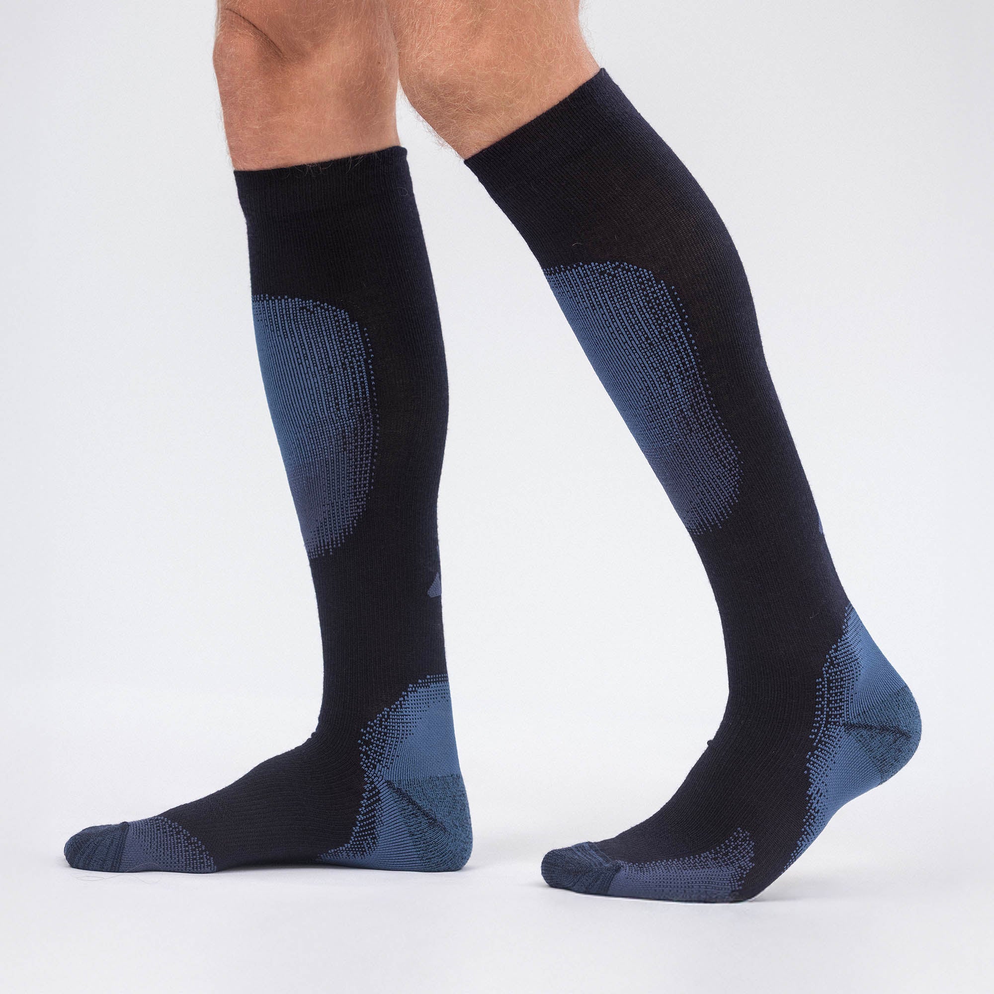 A person wears knee-high, black Socks Merino – Alpine with blue cushioned zones, standing on a white background. Only their lower legs and feet are visible, ideal for alpine skiing or pairing with ski boots.