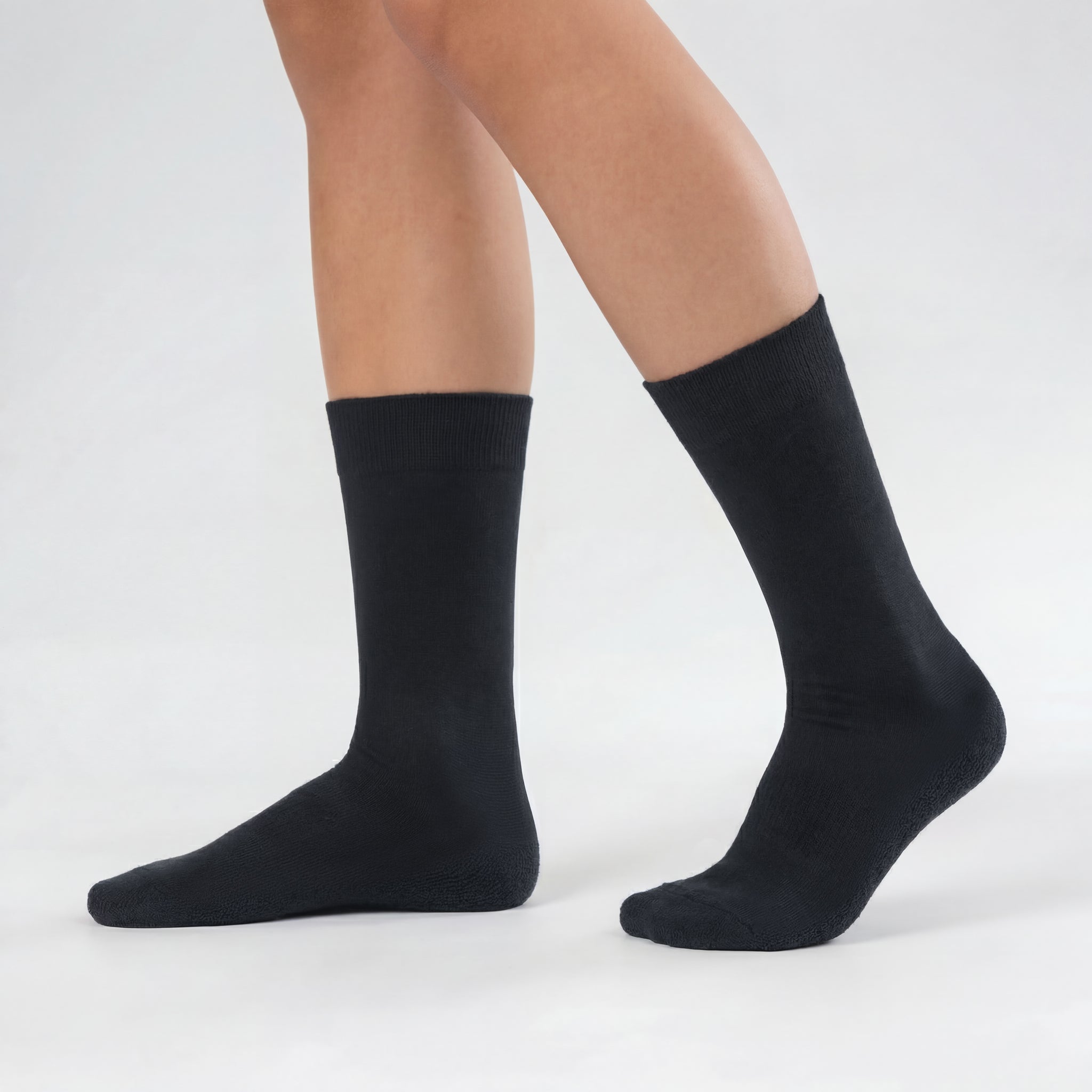 A person wears black Socks 3-pack – Daily Medium with a terry sole, standing on a light gray surface, one foot flat and the other slightly raised at the heel.