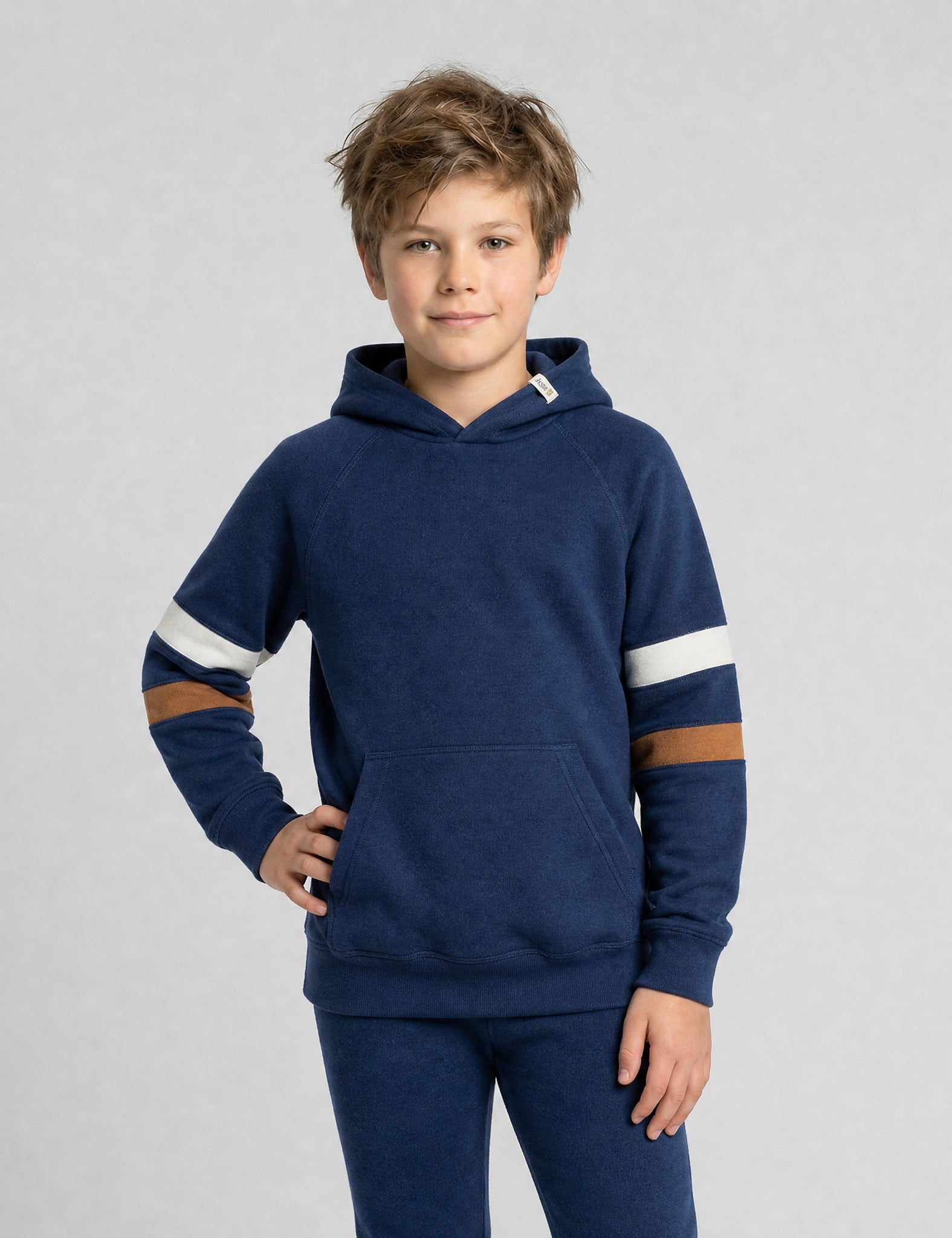 A young boy with light brown hair models the Hoodie merino - Janus Go 250 Kids, a children’s hoodie with white and brown striped sleeves and matching blue pants, standing with one hand on his hip against a plain light gray background.