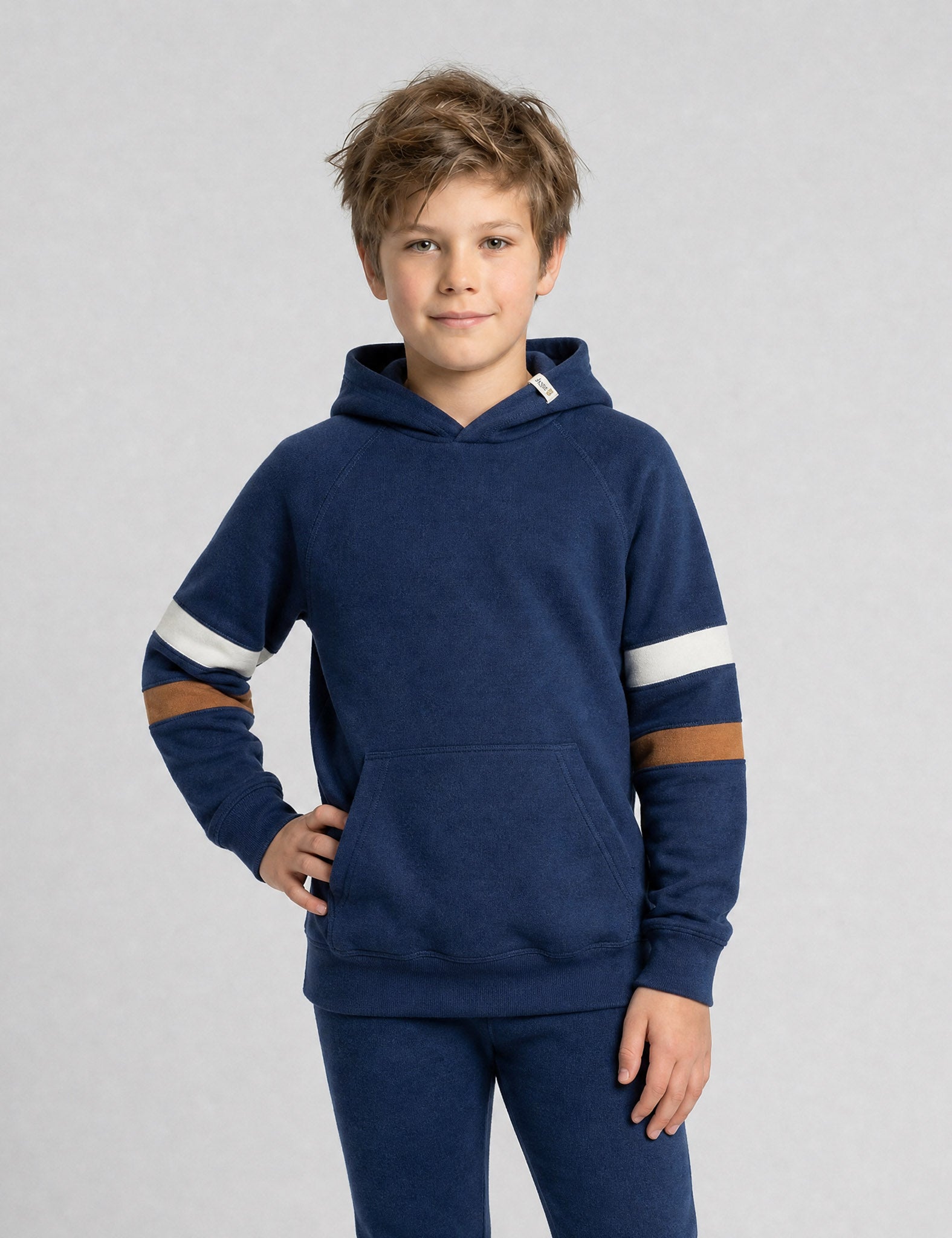 A young boy with light brown hair models the Hoodie merino - Janus Go 250 Kids, a children’s hoodie with white and brown striped sleeves and matching blue pants, standing with one hand on his hip against a plain light gray background.
