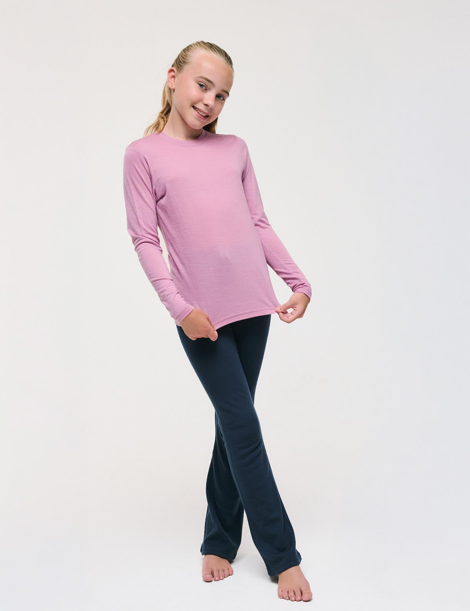 A young girl stands barefoot on a white background, smiling as she holds the sides of her Crewneck – Merino Kids Light shirt to showcase its soft texture. She wears the long-sleeve pink merino top paired with dark blue pants.