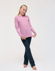 A young girl stands barefoot on a white background, smiling as she holds the sides of her Crewneck – Merino Kids Light shirt to showcase its soft texture. She wears the long-sleeve pink merino top paired with dark blue pants.