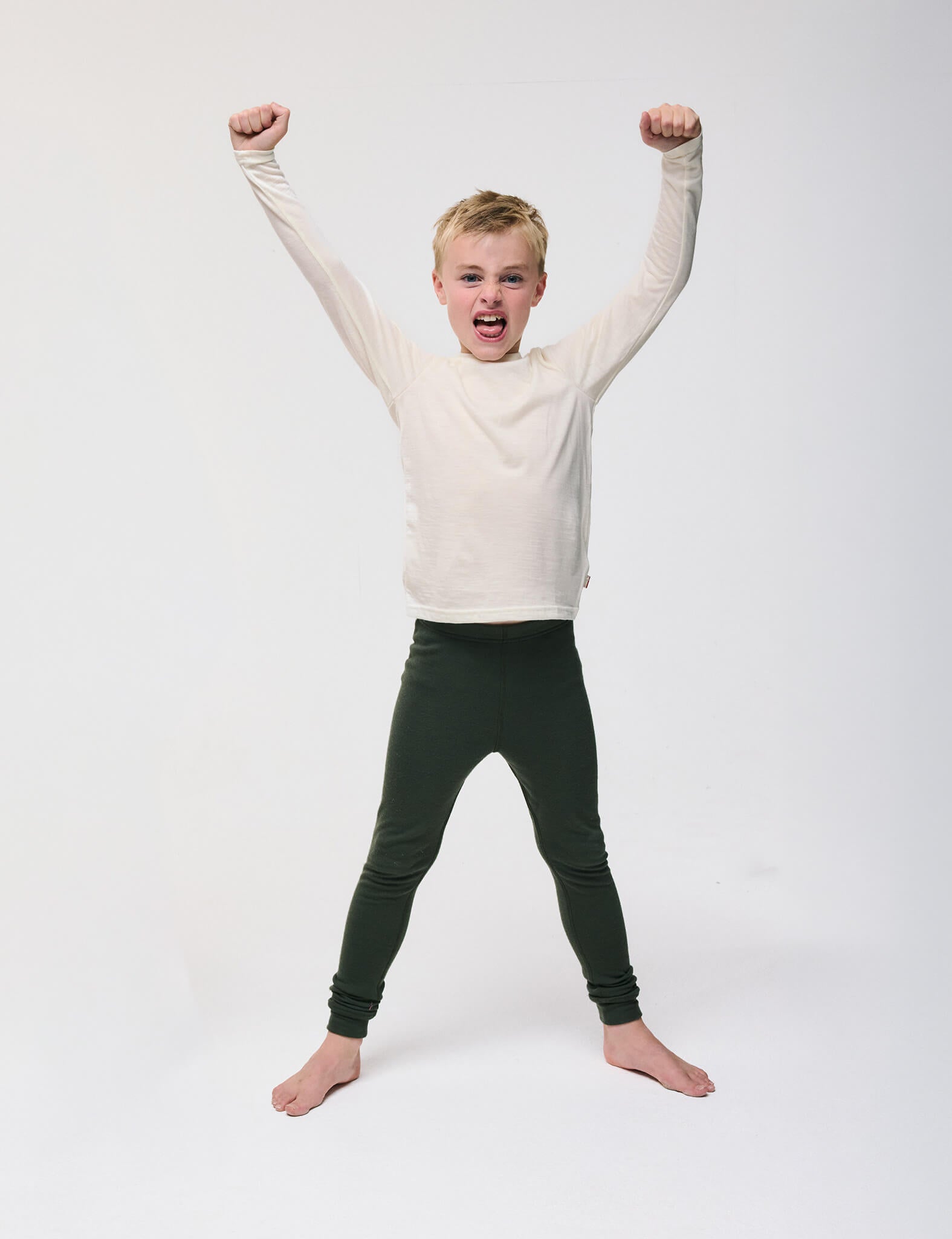 A young boy stands barefoot with legs apart, wearing the Crewneck – Merino Kids Light in cream and dark pants, raising both arms triumphantly and smiling against a plain white background.