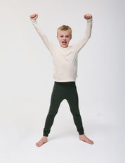 A young boy stands barefoot with legs apart, wearing the Crewneck – Merino Kids Light in cream and dark pants, raising both arms triumphantly and smiling against a plain white background.