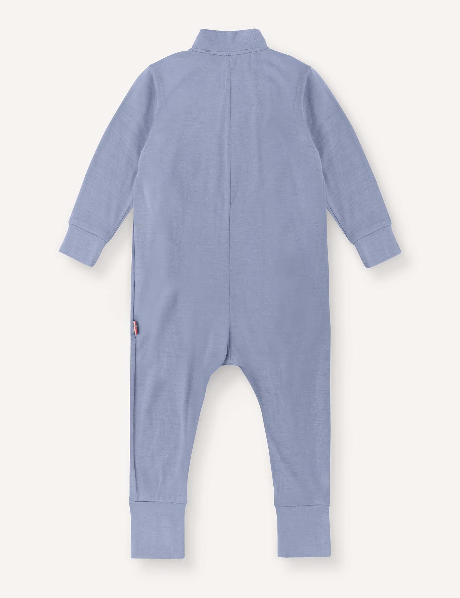 The Pyjamas – Merino Baby Light is a long-sleeve, light blue onesie shown from the back. Designed as breathable pyjamas, it features ribbed cuffs on the sleeves and legs and is finished with a small logo tag on the left side near the hip.