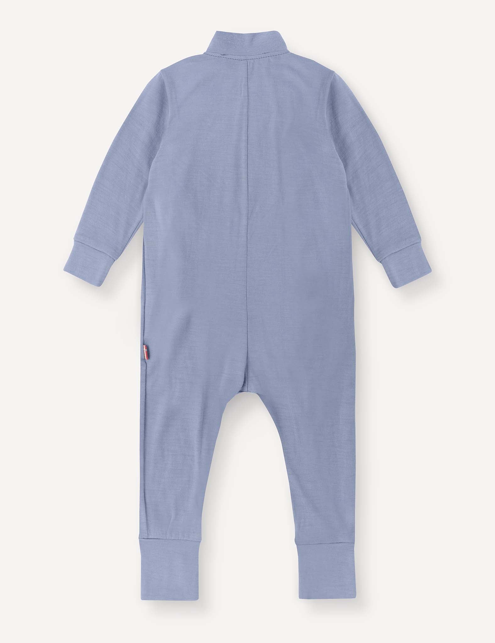 The Pyjamas – Merino Baby Light is a long-sleeve, light blue onesie shown from the back. Designed as breathable pyjamas, it features ribbed cuffs on the sleeves and legs and is finished with a small logo tag on the left side near the hip.