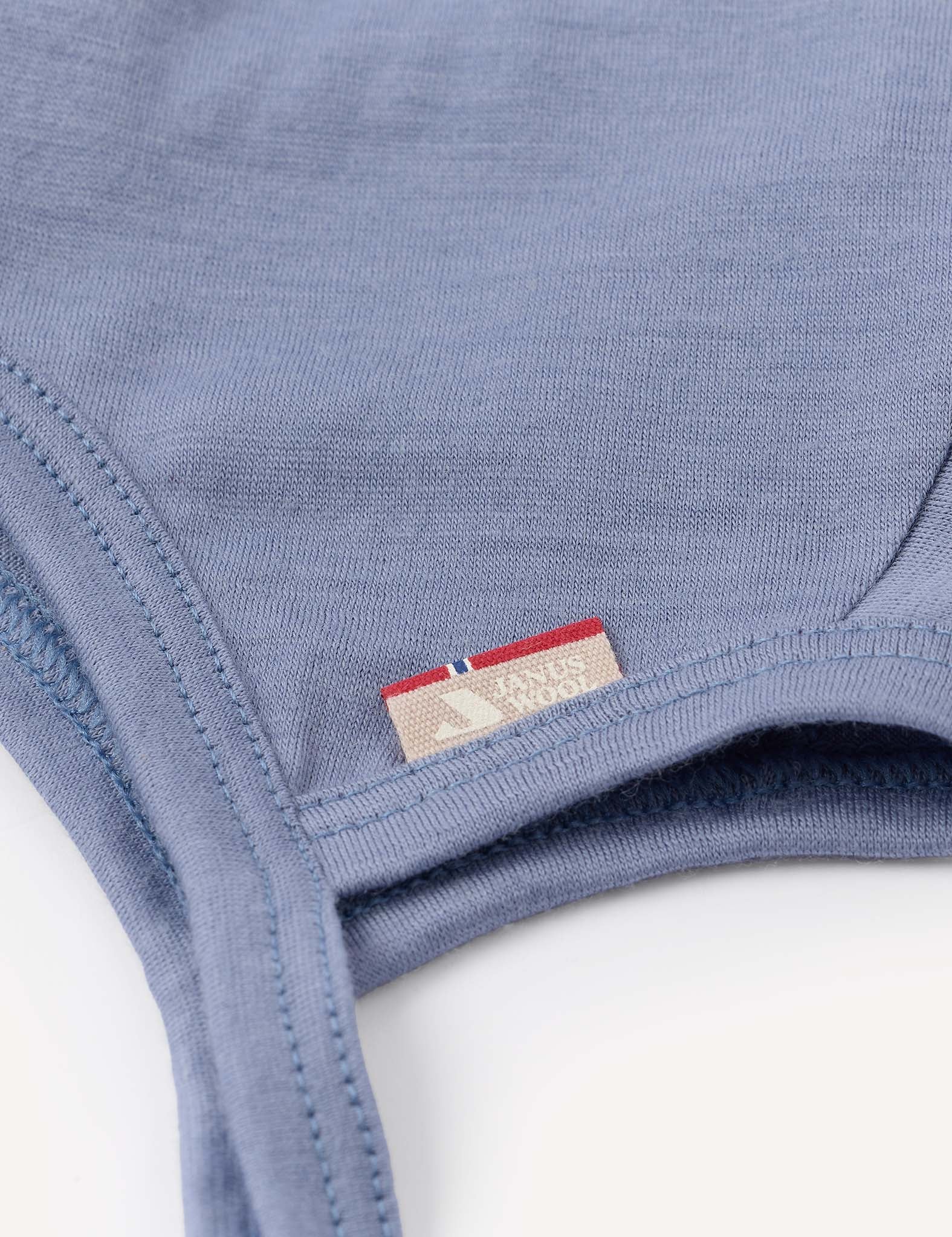 Close-up of the Bonnet – Merino Baby Light in blue, showcasing its soft, smooth merino fabric with visible stitching and a small beige tag featuring red and blue stripes along with text. The material appears breathable and gentle, ideal for a baby bonnet.
