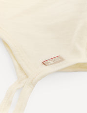 Close-up of the Bonnet – Merino Baby Light in a cream shade, showing a small fabric tag with text and a red accent. The soft, lightweight merino wool fabric is similar to materials used in breathable baby hats and bonnets.