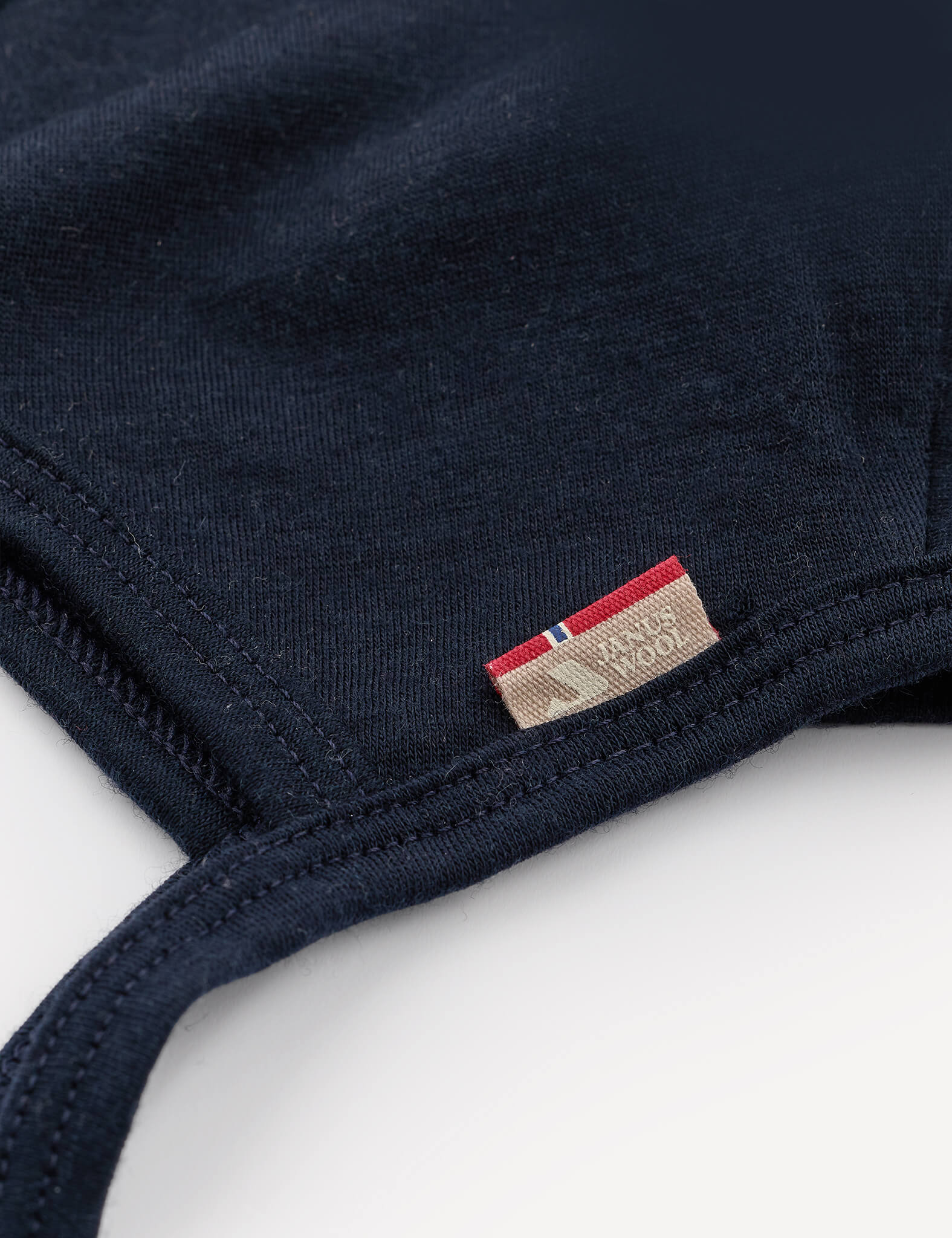 Close-up of the Bonnet – Merino Baby Light in navy blue, featuring a small fabric tag with red, beige, and blue accents. The partially visible tag is sewn into the seam of this lightweight bonnet, which is crafted from soft materials.