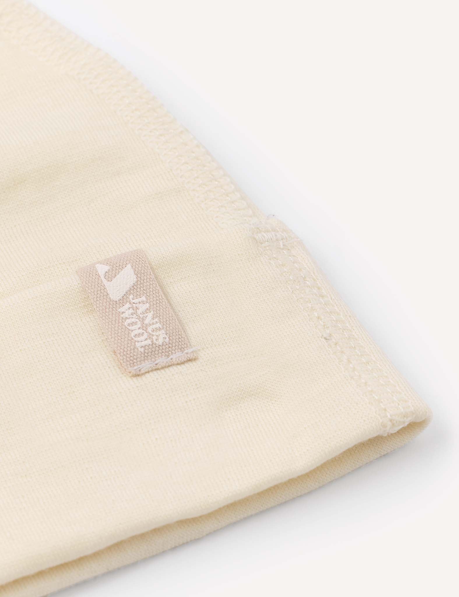 A close-up of the Beanie – Merino Baby Light in beige shows a small rectangular tag sewn onto it, featuring the words “pure born” and a white logo, with visible stitching along the edge.