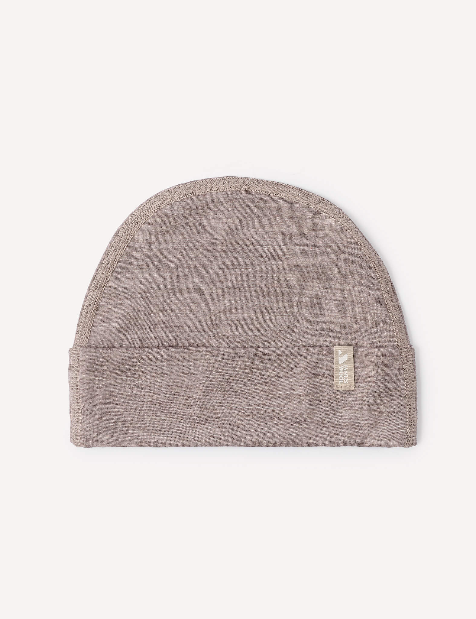 A lightweight, light brown Beanie – Merino Baby Light lies flat on a white background. A small beige fabric tag featuring white text and a logo is sewn onto the folded edge of the merino wool beanie.