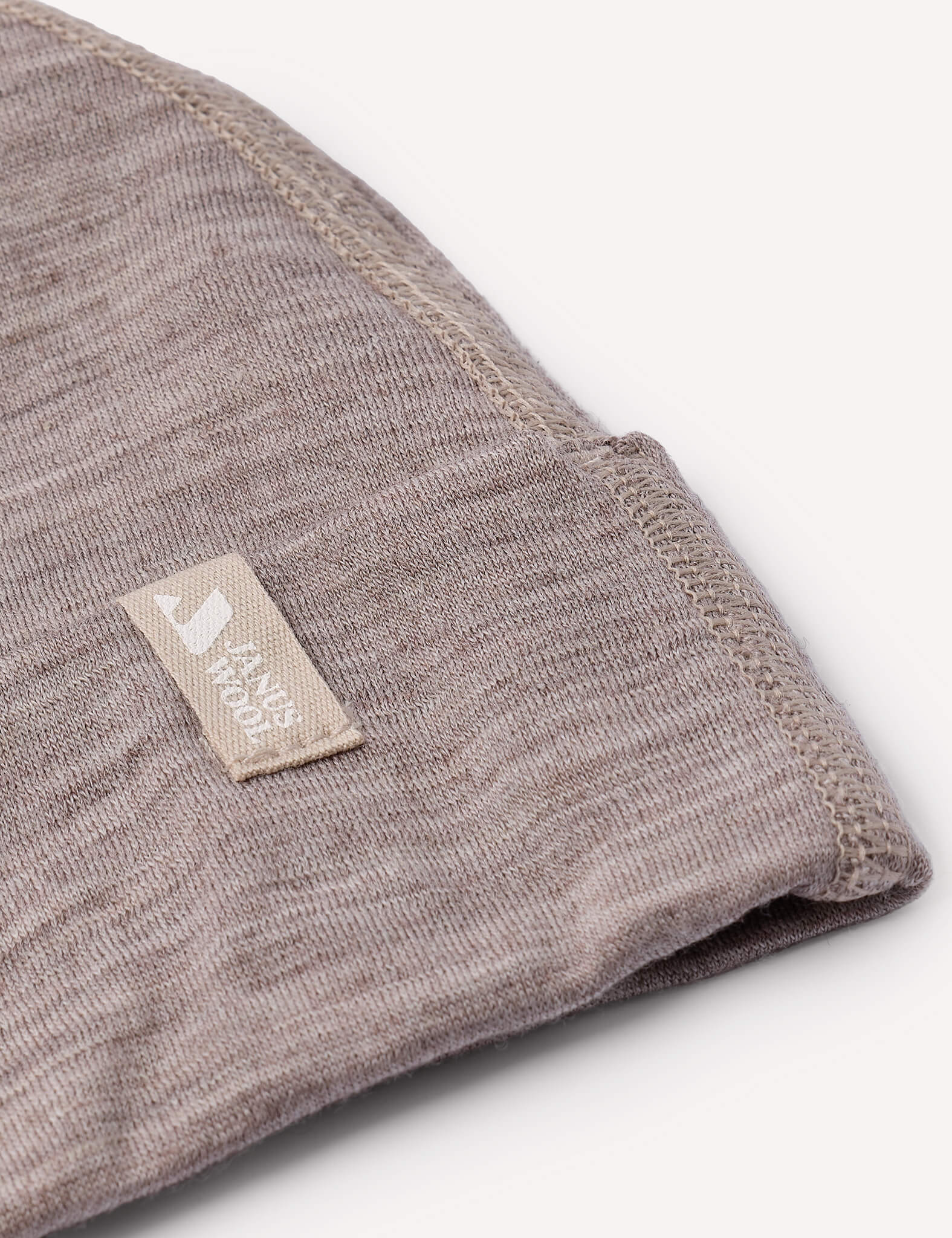 A close-up of the Beanie – Merino Baby Light in a folded brownish-grey merino wool fabric, featuring visible stitching and a small beige tag that reads SANDY NOON, highlighting its lightweight texture.