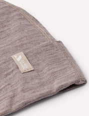 A close-up of the Beanie – Merino Baby Light in a folded brownish-grey merino wool fabric, featuring visible stitching and a small beige tag that reads SANDY NOON, highlighting its lightweight texture.