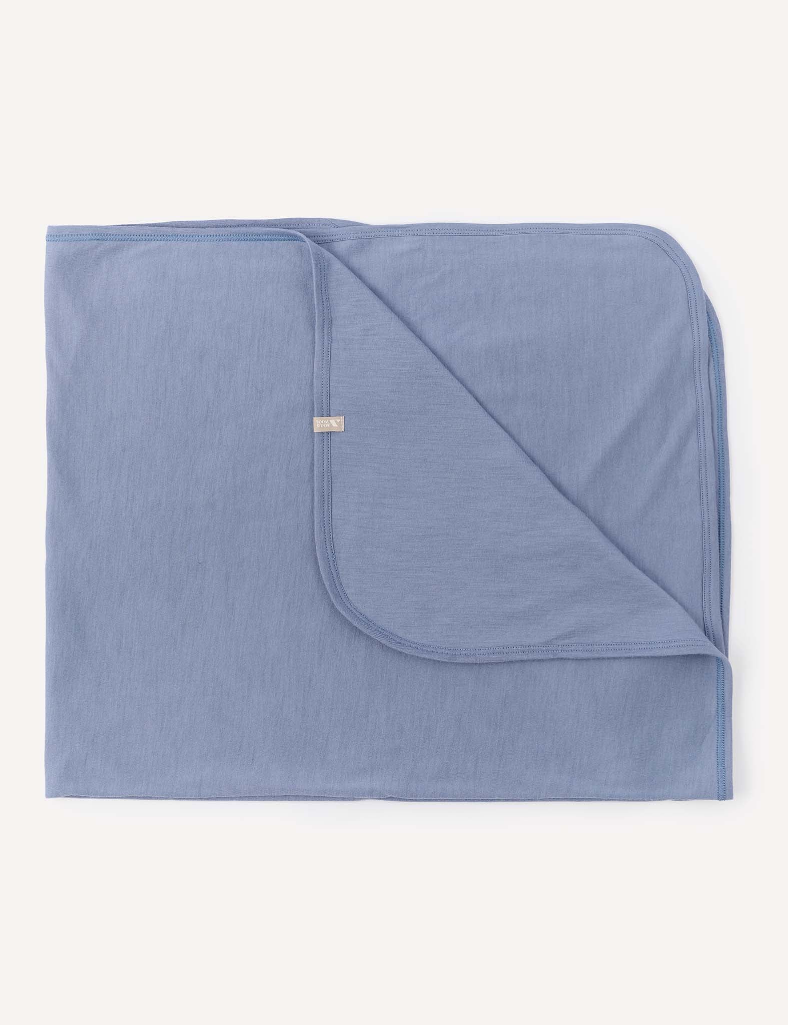 A neatly folded Blanket – Merino Baby Light in Troposphere, with one corner partially turned over to reveal the underside. The blanket features rounded edges and a small fabric tag attached near the turned corner.