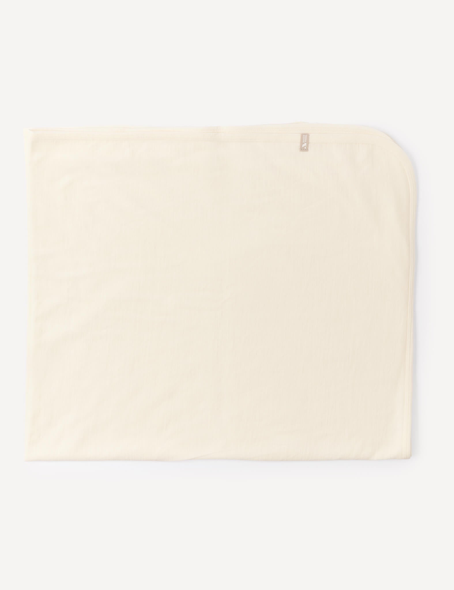 A cream-colored, rectangular Blanket – Merino Baby Light, designed as a lightweight baby blanket with a small tag in one corner, is laid flat on a white background.