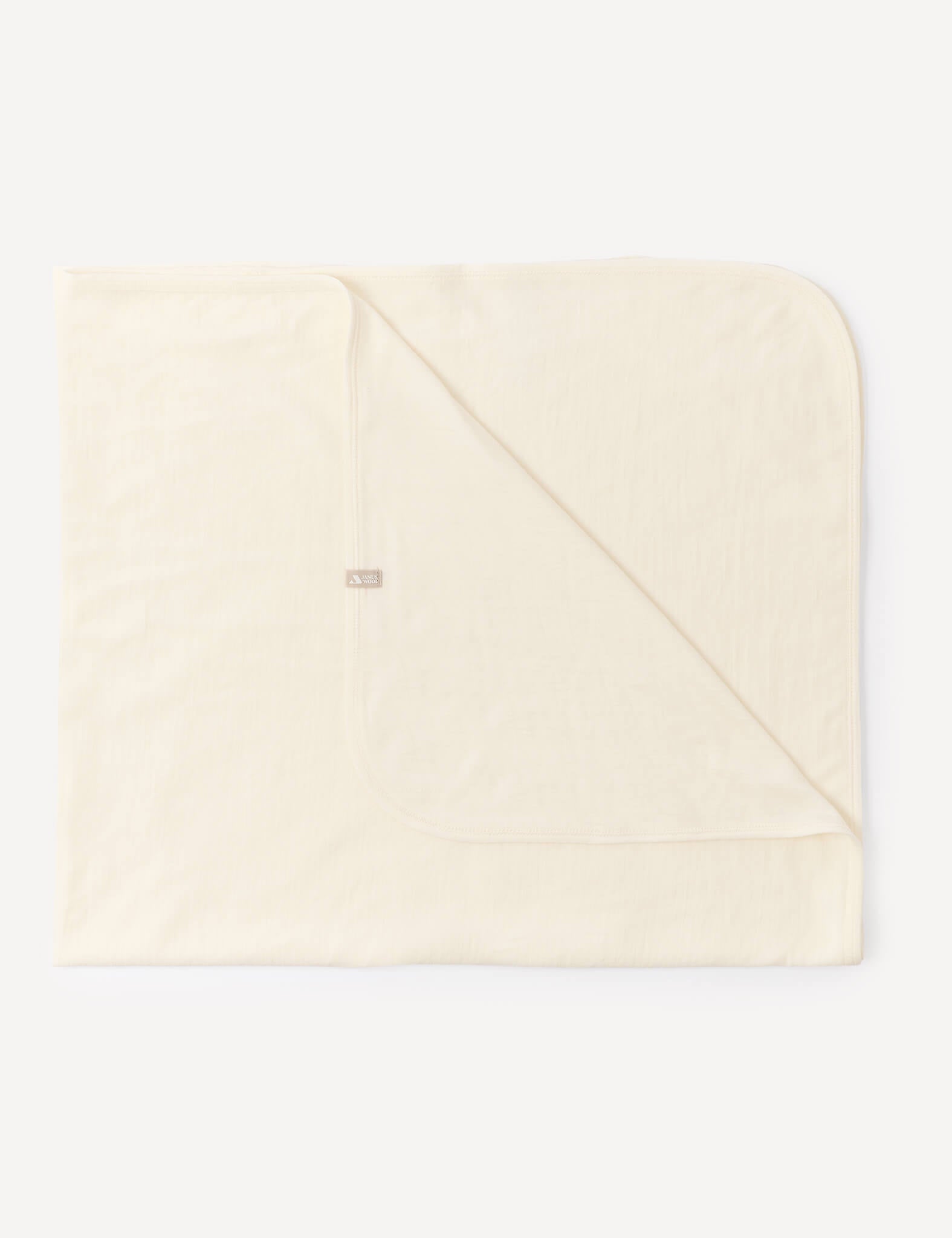 A marshmallow-colored, square Blanket – Merino Baby Light is laid flat with one corner folded over, showcasing its soft, breathable fabric and a small, rectangular tag sewn near the edge.