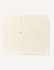 A marshmallow-colored, square Blanket – Merino Baby Light is laid flat with one corner folded over, showcasing its soft, breathable fabric and a small, rectangular tag sewn near the edge.