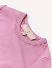 A close-up of the collar and beige fabric tags of the Body – Merino Baby Light, a light purple baby bodysuit, displayed on a white background.