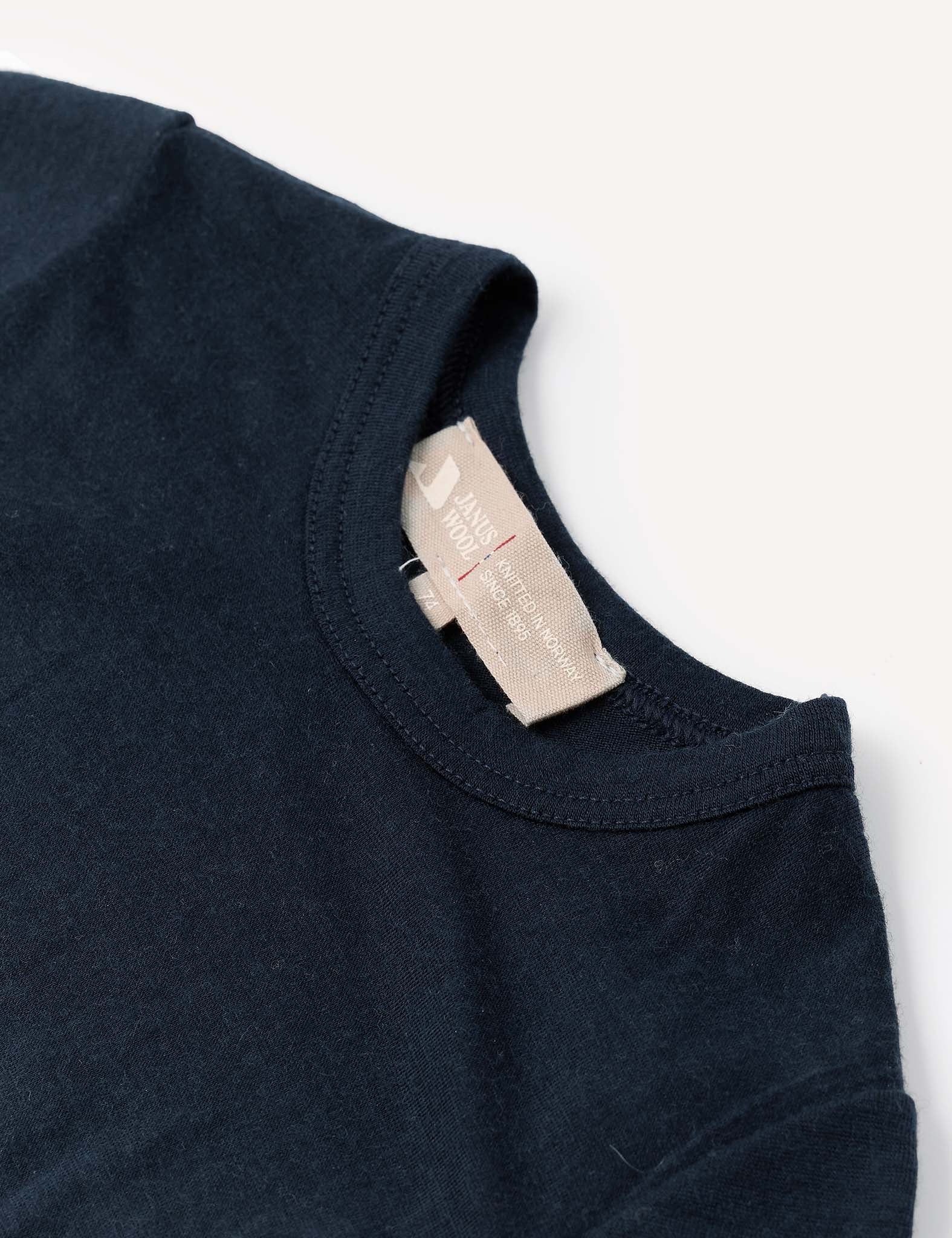 A close-up of the Body – Merino Baby Light in navy blue, highlighting the crewneck collar and a beige fabric tag with text, set against a white background and evoking the feel of airy, breathable baby clothing.
