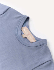 A close-up of the Body – Merino Baby Light in light blue displays the neckline and a beige brand tag sewn inside the collar. The merino wool bodysuit is laid flat on a white surface.