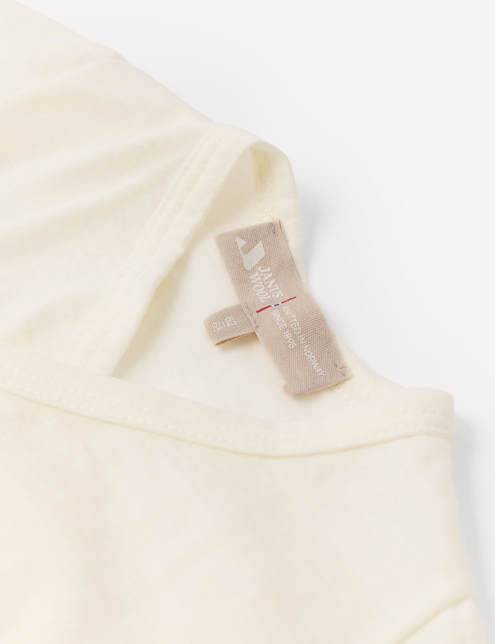 A close-up of a cream-colored shirt’s collar and beige tag, displaying size and care information on a white background, highlights the quality of the Wrapover Bodysuit – Merino Baby Light.