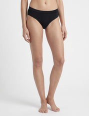 A person stands barefoot on a white background, wearing Briefs – Classic Merino 160 Women, known for their breathable, temperature-regulating qualities. The person's face is not shown in the image.