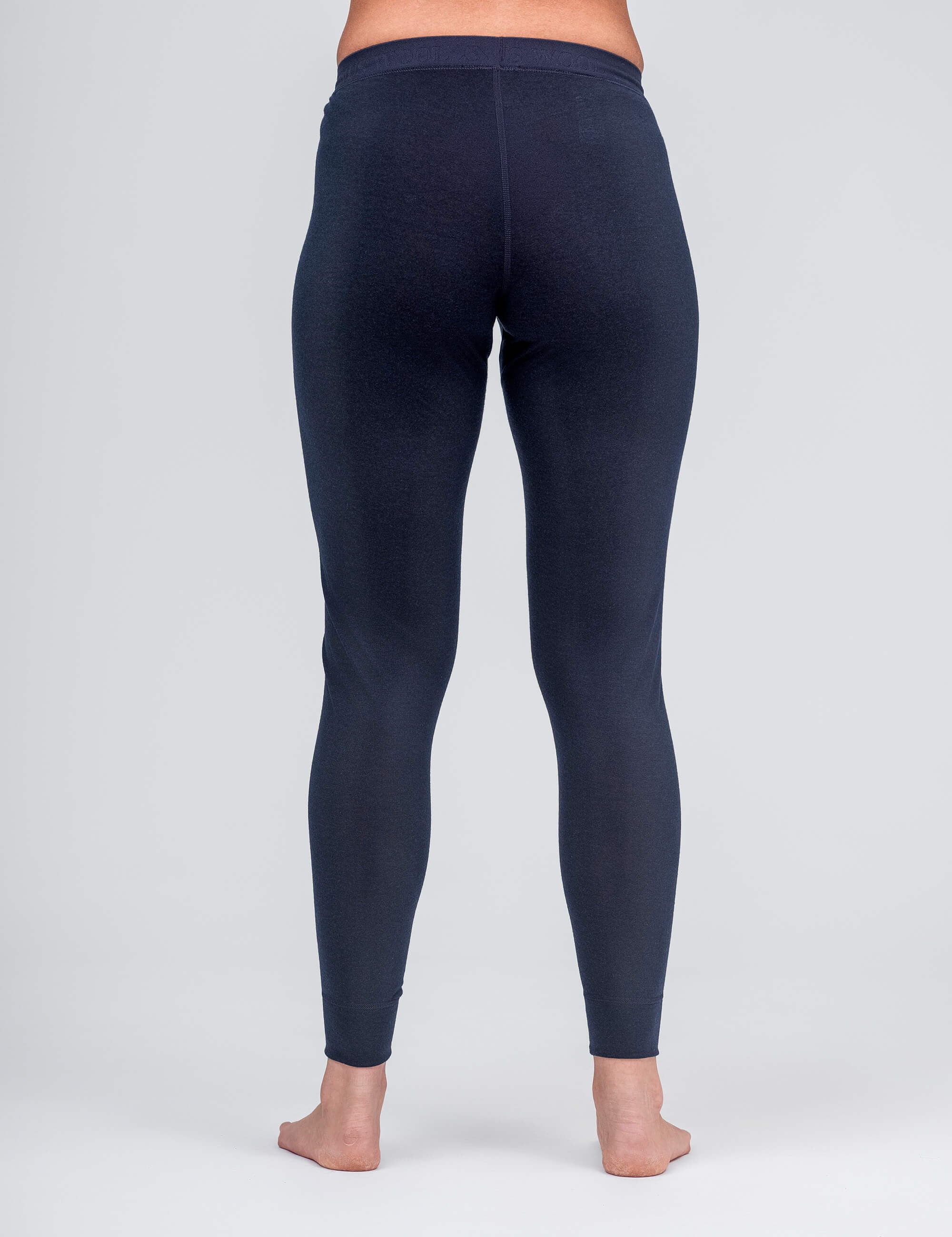 A person stands barefoot, facing away from the camera, wearing Classic Merino 160 Women leggings in dark blue, shown from waist to ankles against a plain light grey background.