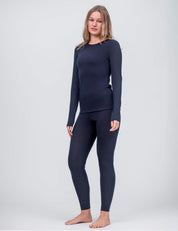 A woman, barefoot against a plain light background, wears the Classic Merino 160 Women leggings in navy blue with a matching long-sleeve top, standing slightly turned to the side with a neutral expression.