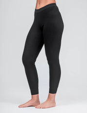 A person stands barefoot on a plain background, wearing Classic Merino 160 Women Leggings in black. Cropped from midriff to feet, the image shows the breathable, temperature-regulating fit from the side and front.