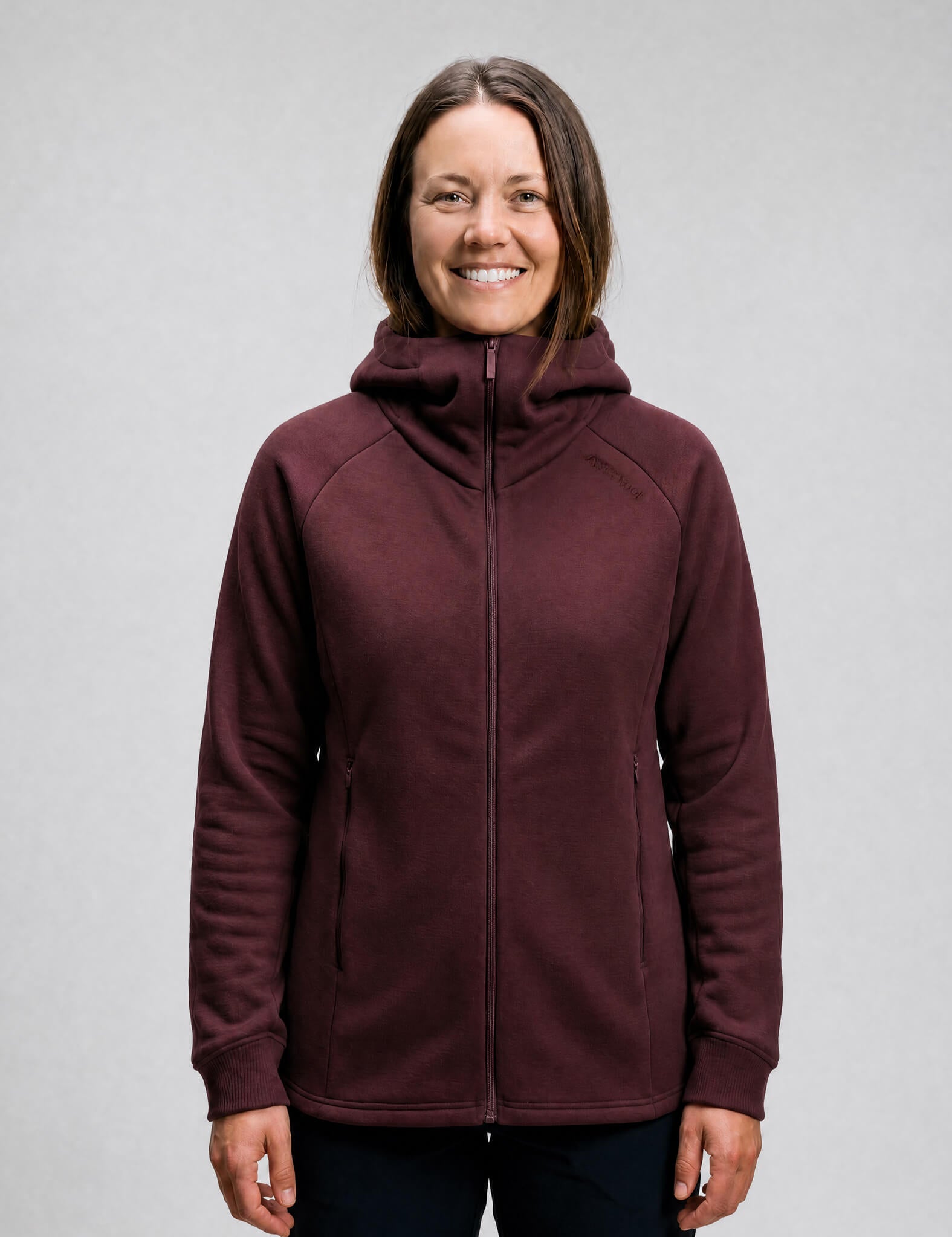 A woman with straight brown hair wears the Hooded Jacket Merino – AirWool 280 Women in maroon, standing and smiling at the camera against a plain light gray background.