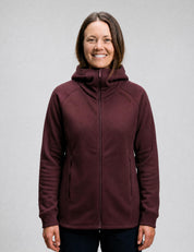 A woman with straight brown hair wears the Hooded Jacket Merino – AirWool 280 Women in maroon, standing and smiling at the camera against a plain light gray background.