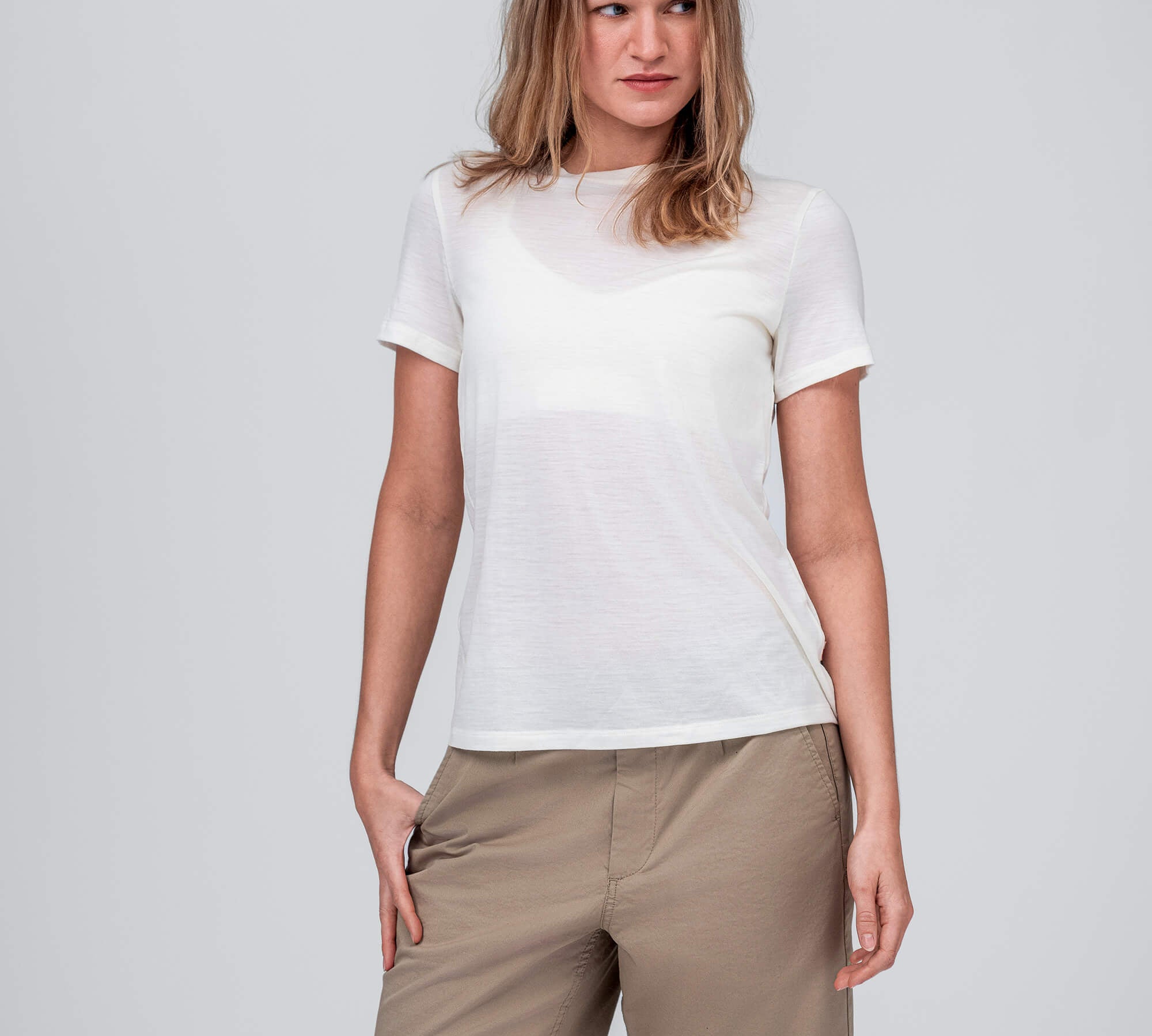 A woman stands against a light gray background, wearing the T-shirt Regular – Classic Merino 160 Women in Marshmallow and light brown knee-length shorts. She looks slightly to her right, with one hand resting in her pocket.