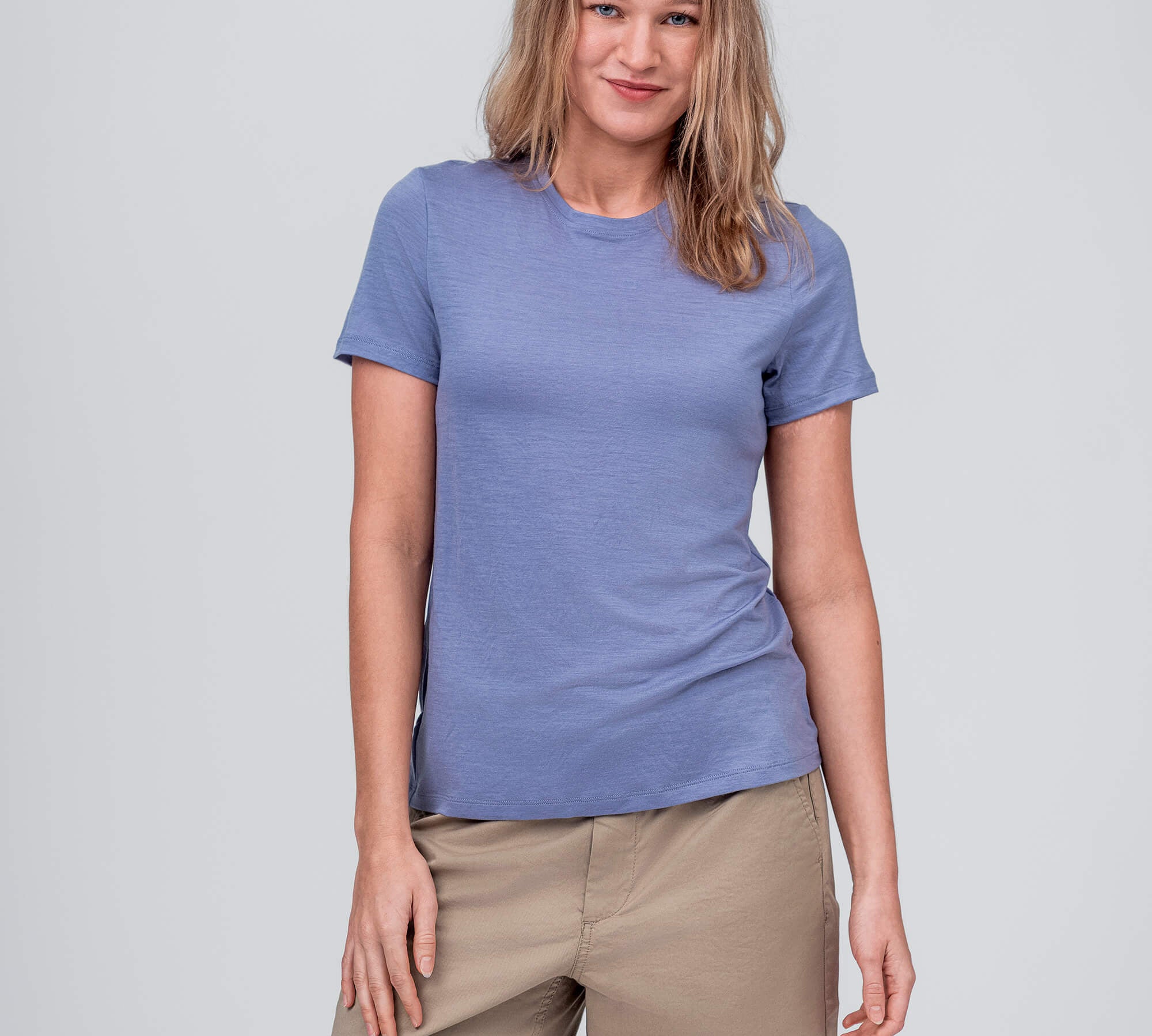 A woman with light brown hair wears the T-shirt Regular – Classic Merino 160 Women in Troposphere and beige shorts, standing against a plain light gray background, smiling gently with one hand relaxed by her side.