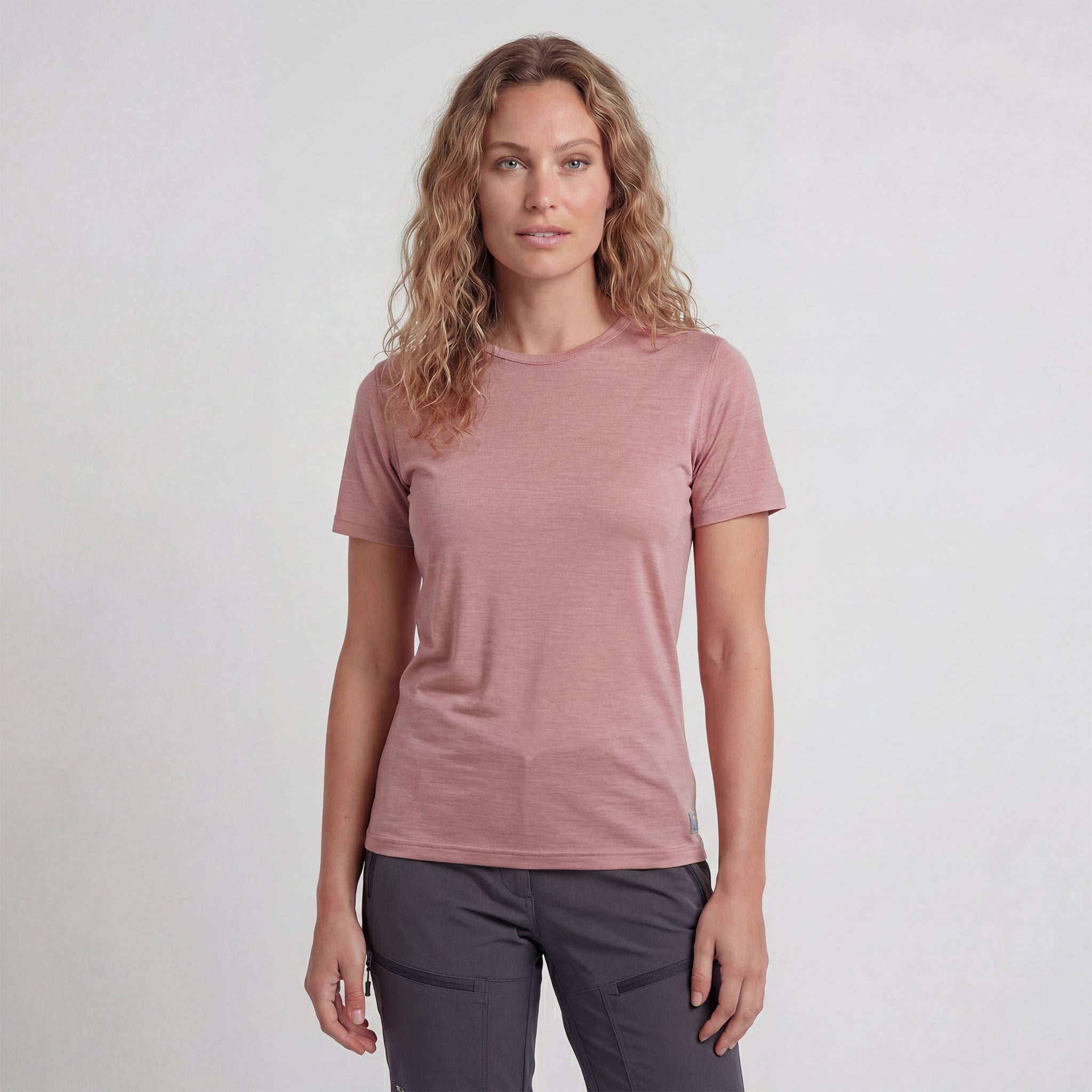 A woman with wavy blonde hair wears a T-shirt merino - Lightwool 160 Women in light mauve and dark gray pants, standing facing forward against a plain light background.