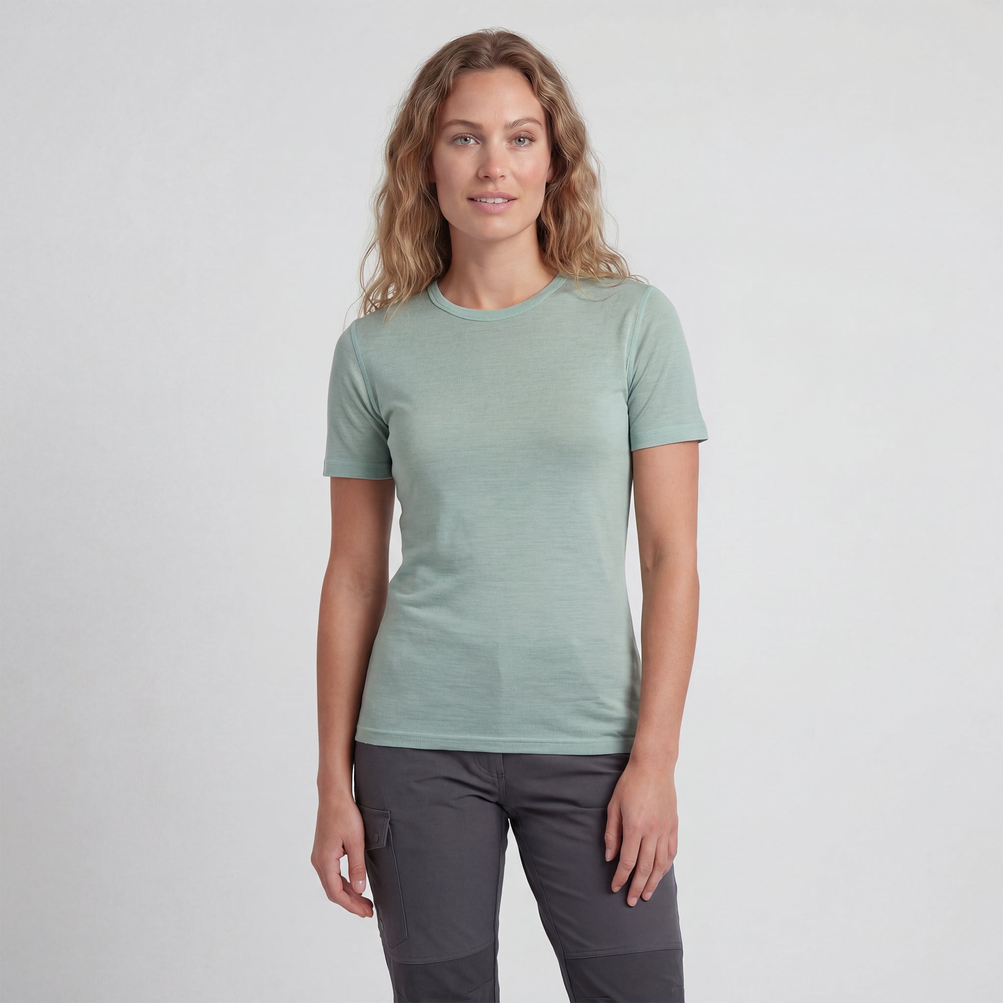 A woman with wavy blonde hair wears a T-shirt merino - Lightwool 160 Women in turquoise and dark gray pants, standing before a plain light background and smiling softly at the camera.
