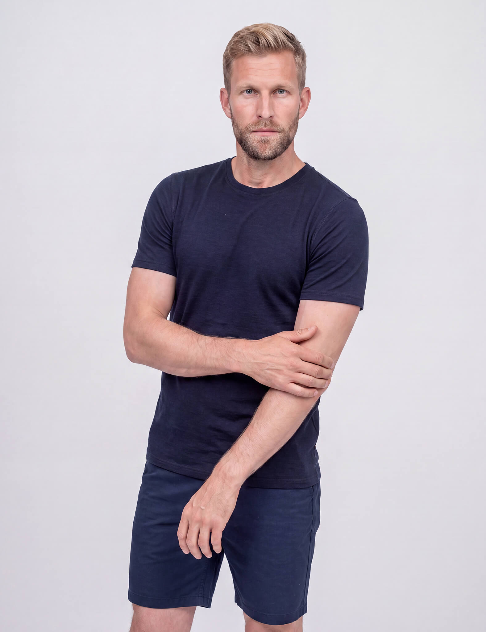 A man with short, tousled hair and a beard stands against a plain background, wearing the T-shirt Active – Classic Merino 160 Men in Navy, paired with matching shorts, looking directly at the camera with a neutral expression.