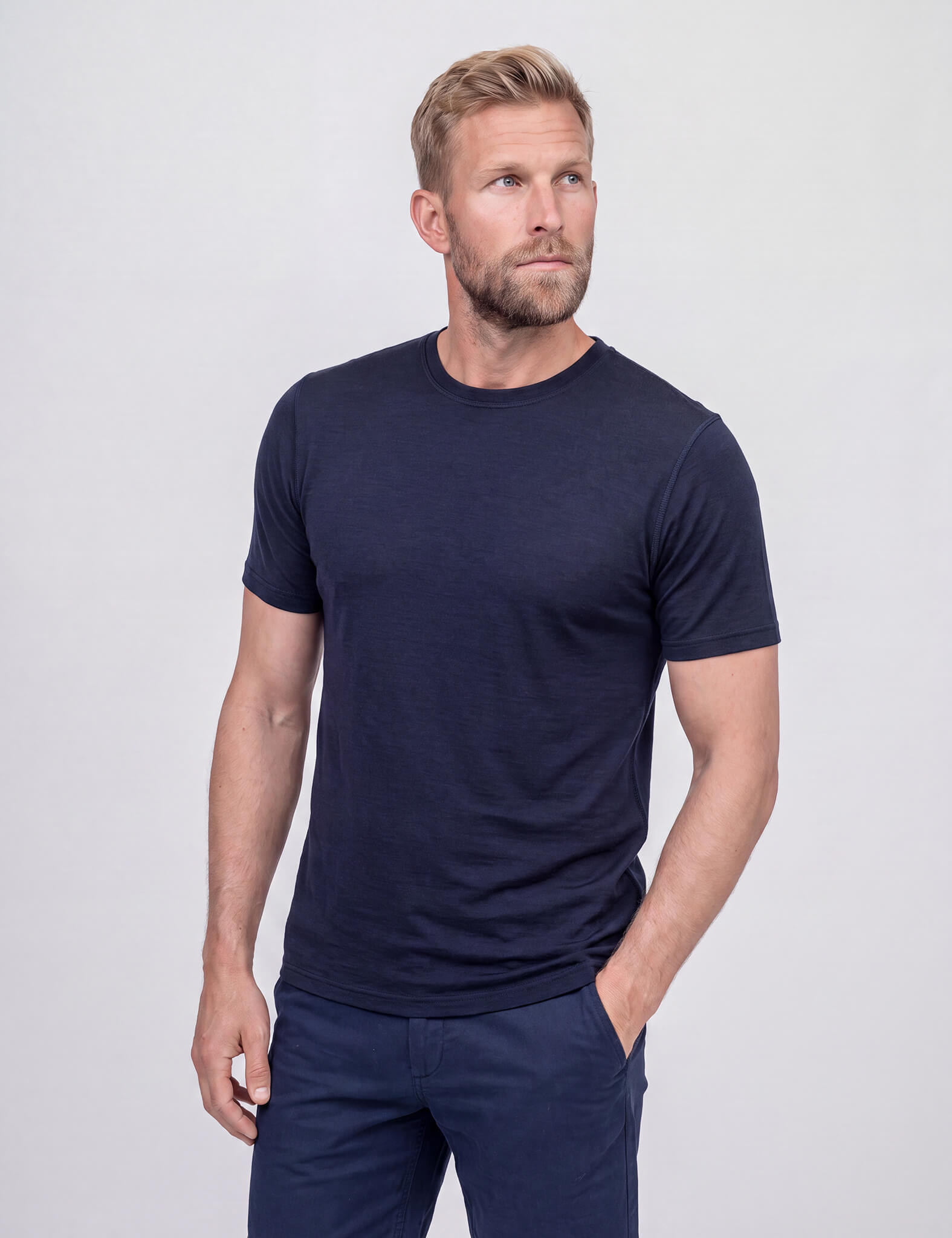 A man with short hair and a beard wears the T-shirt Active – Classic Merino 160 Men, paired with matching shorts, as he poses with one hand in his pocket against a plain light gray background.