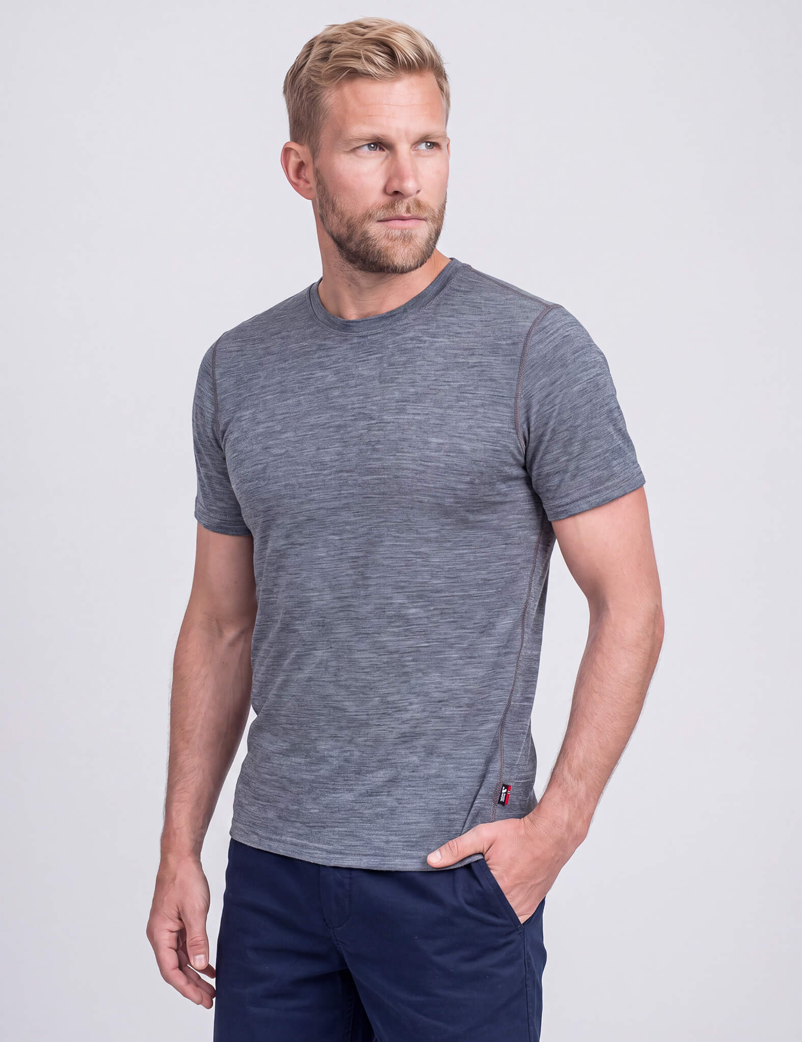 A bearded man stands against a plain light gray background, wearing a grey melange T-shirt Active – Classic Merino 160 Men and navy shorts, looking to his left with one hand in his pocket.