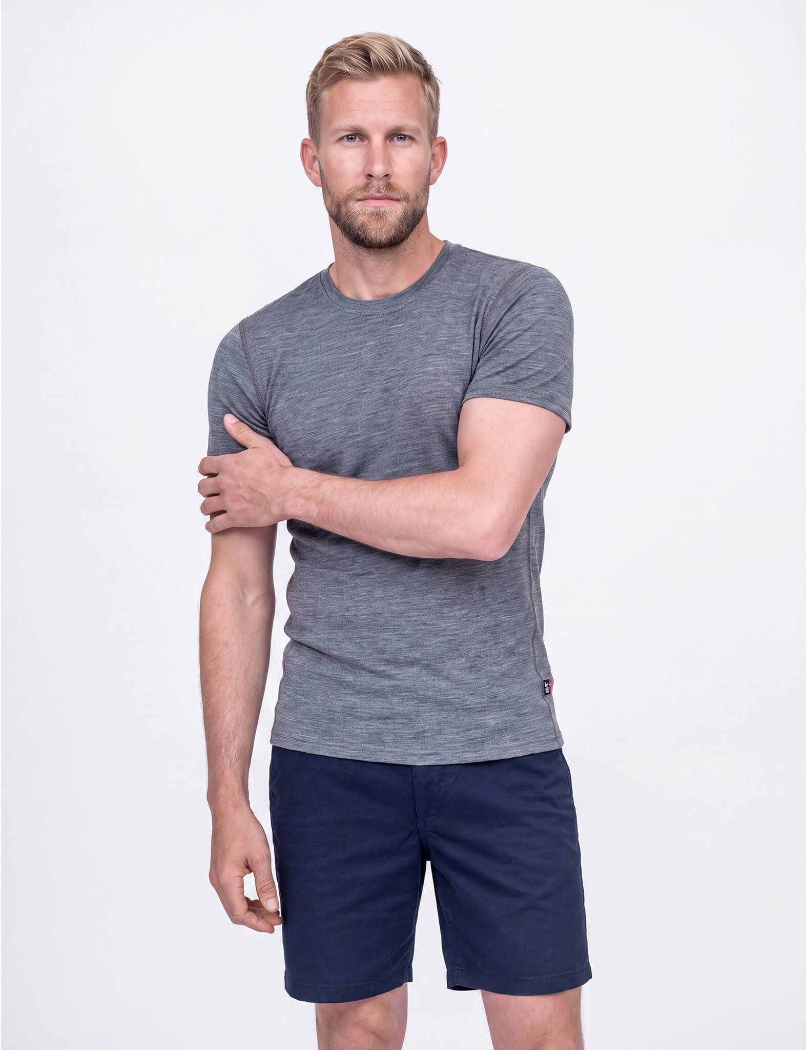 A bearded man wearing the T-shirt Active – Classic Merino 160 Men and navy shorts looks down as he inspects his upper left arm with his right hand against a plain, light background.