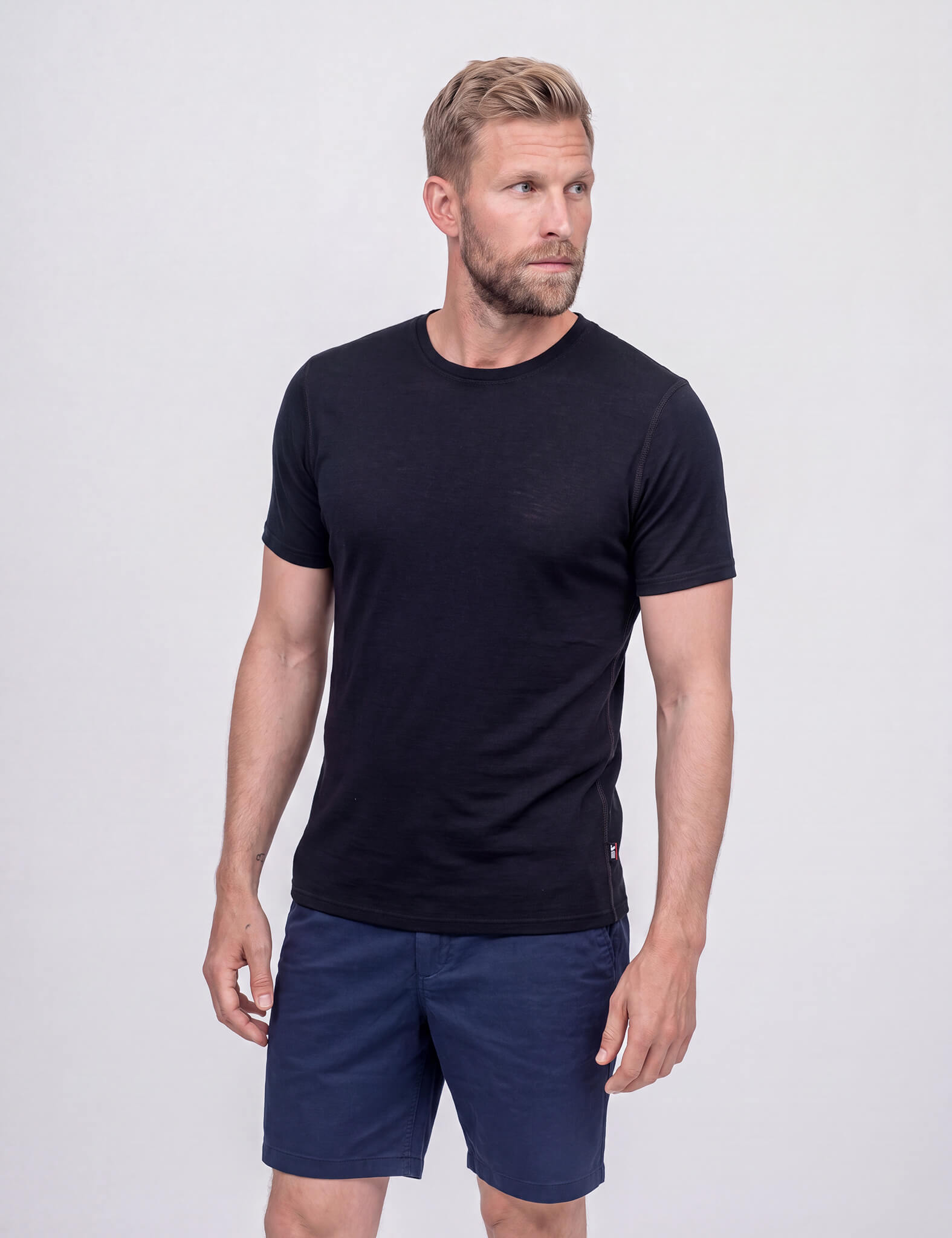 A light-skinned man with a beard and short, tousled hair wears the T-shirt Active – Classic Merino 160 Men in black, known for temperature regulation, paired with dark blue pants. He stands against a plain white background looking slightly left.