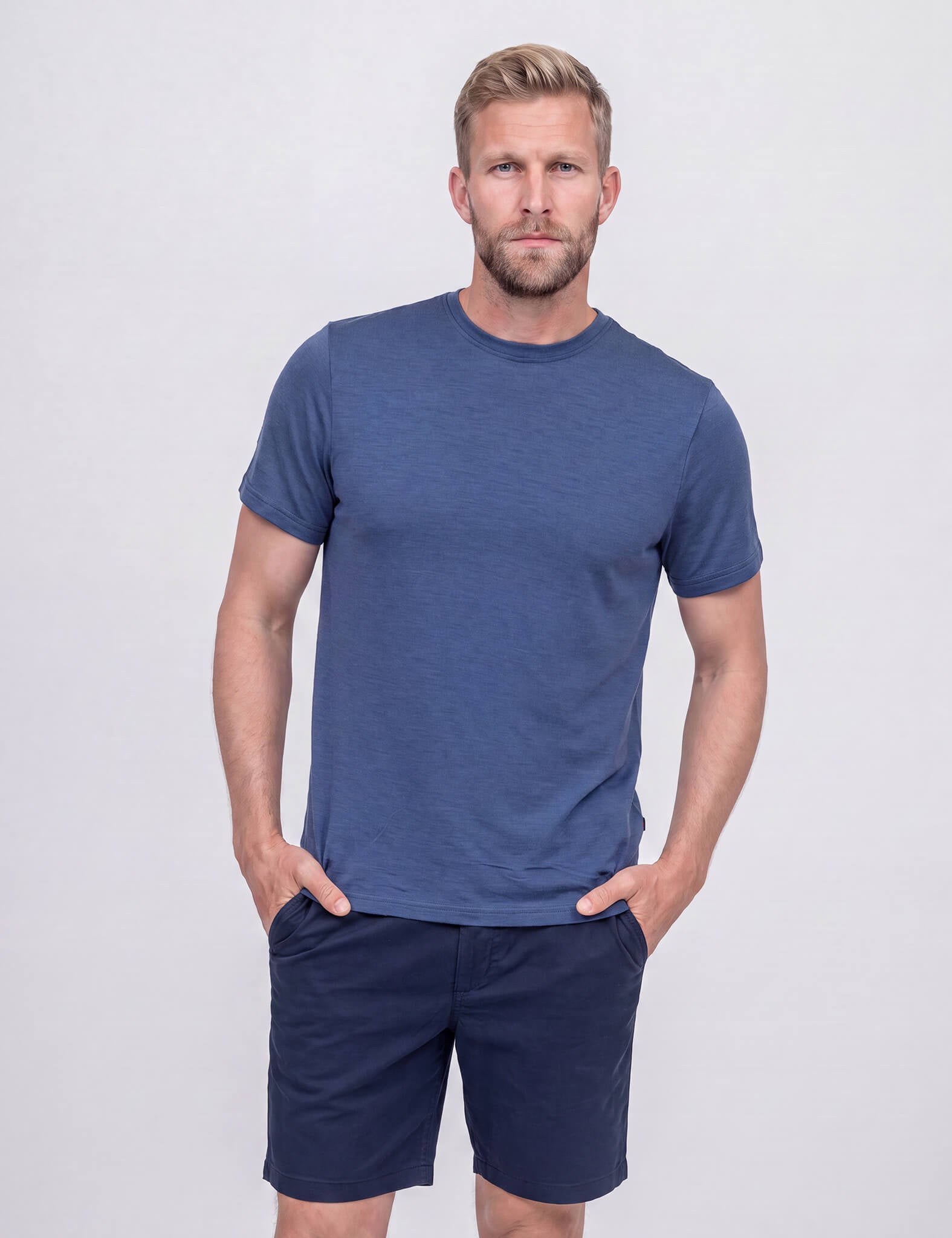 A man with short brown hair and a beard stands against a light gray background, wearing the T-shirt Regular – Classic Merino 160 Men in Ensign Blue, paired with dark blue shorts. His hands are in his pockets and he has a slight smile.