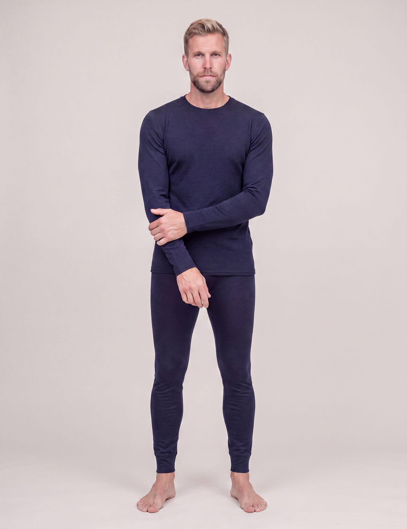 A man stands barefoot against a plain background, wearing the Crew Neck – Classic Merino 160 Men long-sleeve layer and matching fitted pants, with one hand resting on his opposite arm.