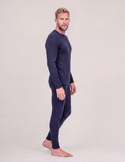 A man with short blond hair stands barefoot in profile against a light gray background, wearing the Crew Neck – Classic Merino 160 Men top in navy with matching leggings—an ideal travel-friendly layer.